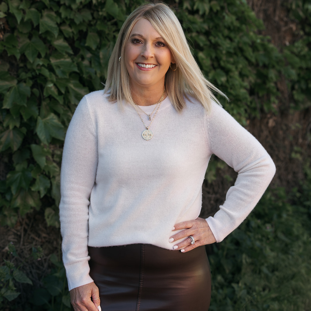 Woman wearing a light gray sweater and dark pants standing against a green leafy background